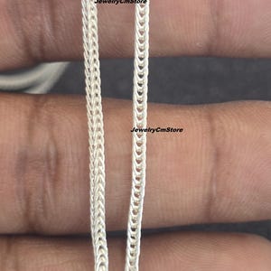Puede incluir: Primer plano de dos collares de cadena plateados. La cadena de la izquierda tiene un diseño tejido, mientras que la cadena de la derecha tiene una serie de eslabones conectados. El texto "JewelryCmStore" es visible en ambas cadenas.