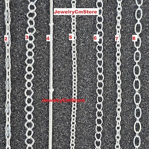 Può includere: Una collezione di nove stili di catene d'argento esposti su uno sfondo scuro e strutturato. Le catene variano nel design, inclusi collegamenti a forma di cuore, ovali e trame a corda. Il testo "JewelryCmStore" è visibile.