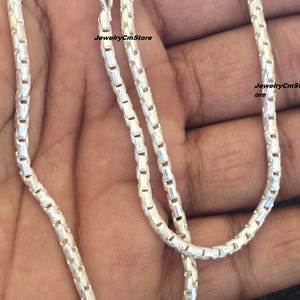 Puede incluir: Un collar plateado con un diseño de cadena de caja. El collar se muestra sobre un fondo neutro. La cadena está hecha de pequeños eslabones rectangulares. El collar tiene un cierre. El texto "JewelryCmStore" es visible.