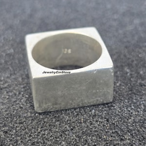 May include: A silver-colored square ring. The ring has a solid, geometric design with a smooth, polished surface. The ring is a minimalist style, suitable for everyday wear. The ring is shown on a dark, textured surface.