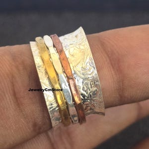 May include: A silver spinner ring with a textured band and floral pattern. It has four smaller bands in gold, white, silver, and copper. The text "JewelryCmStore" is visible on the ring.