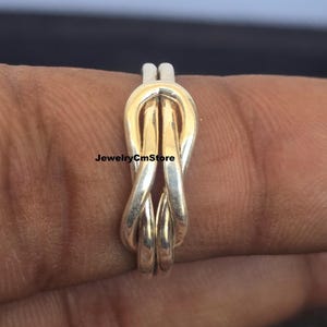 May include: A silver-toned ring with a knot design. The ring features a gold-colored center section. The JewelryCmStore logo is visible on the image. The ring is shown on a finger.