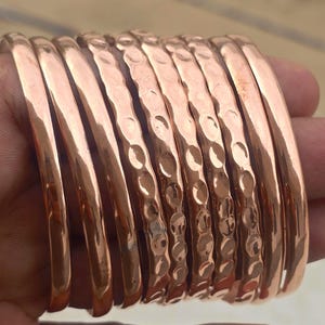Puede incluir: Una colección de brazaletes de cobre. Algunos brazaletes tienen un acabado liso y pulido, mientras que otros presentan una textura martillada. Los brazaletes son de un color marrón rojizo cálido, que refleja la luz.