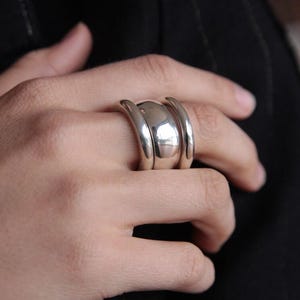 Puede incluir: Un anillo plateado con un centro redondeado y dos bandas curvas a cada lado. El anillo se lleva en un dedo y el metal refleja la luz. El diseño es simple pero audaz.