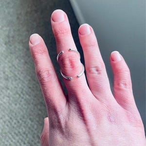 Puede incluir: Un anillo plateado con un diseño abierto, con dos bandas curvas que rodean parcialmente un dedo. El anillo tiene un estilo minimalista con extremos lisos y redondeados.