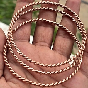 Op de afbeelding: Drie koperkleurige, gedraaide metalen armbanden. De armbanden hebben een doorlopend, spiraalvormig ontwerp. De armbanden worden getoond op een open handpalm, met een vage groene achtergrond.