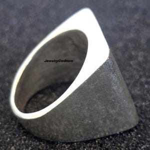 Può includere: Un anello d'argento dal design geometrico unico. L'anello presenta una forma triangolare con una superficie piatta e strutturata e un bordo superiore bianco lucido. Il testo "jewelryCmStore" è visibile sull'anello.