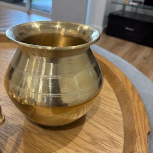 Puede incluir: Una olla de latón dorado con base redondeada y borde acampanado. La olla tiene líneas grabadas horizontales alrededor del cuerpo. La olla está sobre una bandeja de madera.