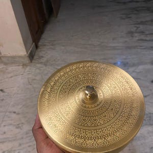 May include: A gold-coloured, round container with an intricate embossed design on the lid. The lid has a small, round knob. The container is held in a hand, showing its size and detail. The design suggests a decorative or storage item.