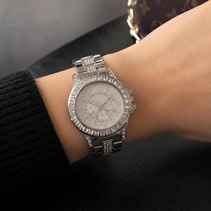 Peut inclure: Une montre-bracelet argentée avec un cadran rond et un bracelet orné de cristaux étincelants. Le cadran de la montre présente un motif texturé et trois sous-cadrans. La montre est portée au poignet, avec une manche noire visible.