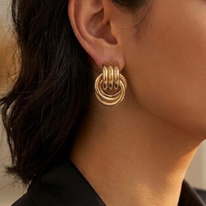 Peut inclure: Une boucle d'oreille créole dorée composée de trois anneaux circulaires lisses et polis, entrelacés et suspendus verticalement au lobe de l'oreille, formant un design superposé et dimensionnel avec une finition métallique réfléchissante.