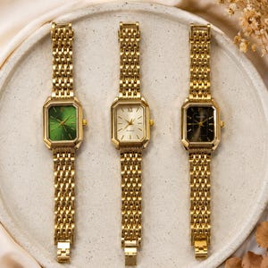 Könnte beinhalten: Drei goldfarbene Uhren mit rechteckigen Zifferblättern und passenden Gliederarmbändern werden auf einem hellen Teller präsentiert. Die Zifferblätter der Uhren sind grün, weiß und schwarz. Die Uhren haben goldfarbene römische Ziffern.