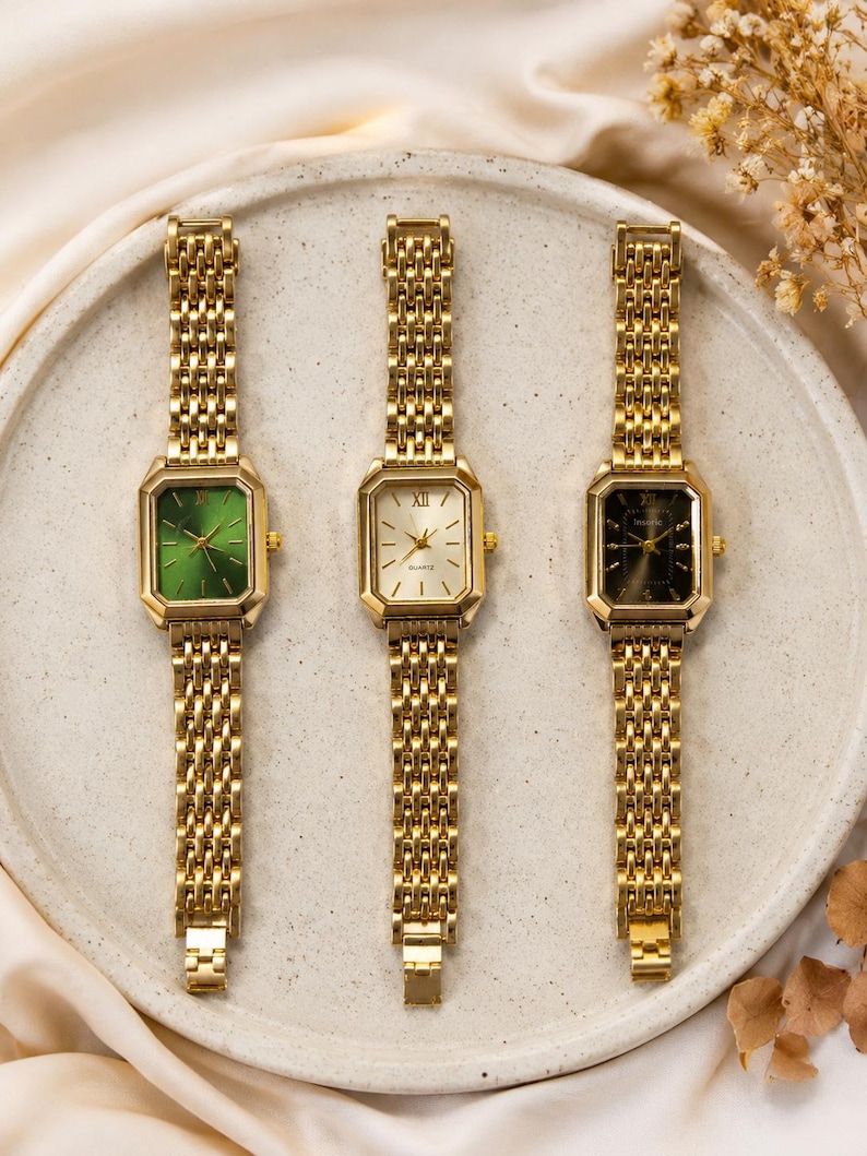 May include: Three gold-toned watches with rectangular faces and matching link bands are displayed on a light-colored plate. The watch faces are green, white, and black. The watches have gold-colored Roman numeral hour markers.