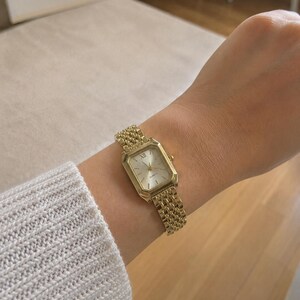May include: A gold-toned wristwatch with a rectangular face and a mesh band. The watch features Roman numeral hour markers and a quartz movement. The band is a woven style, and the watch is worn on a wrist.