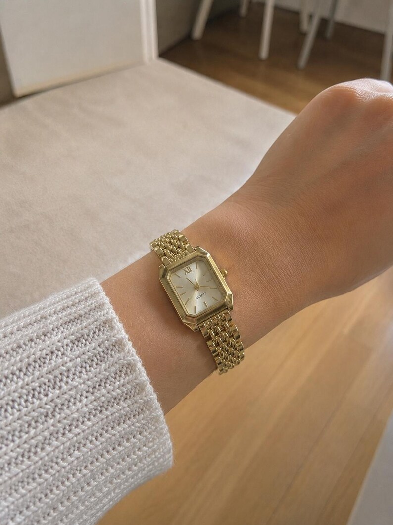 May include: A gold-toned wristwatch with a rectangular face and a mesh band. The watch features Roman numeral hour markers and a quartz movement. The band is a woven style, and the watch is worn on a wrist.