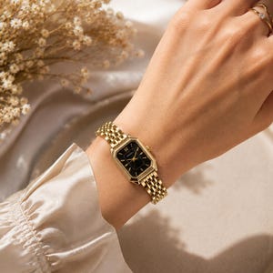 May include: A gold-toned wristwatch with a black face and Roman numeral hour markers is worn on a wrist. The watch has a rectangular face and a woven metal band. A gold ring is on the finger. The background includes a beige silk fabric and dried flowers.