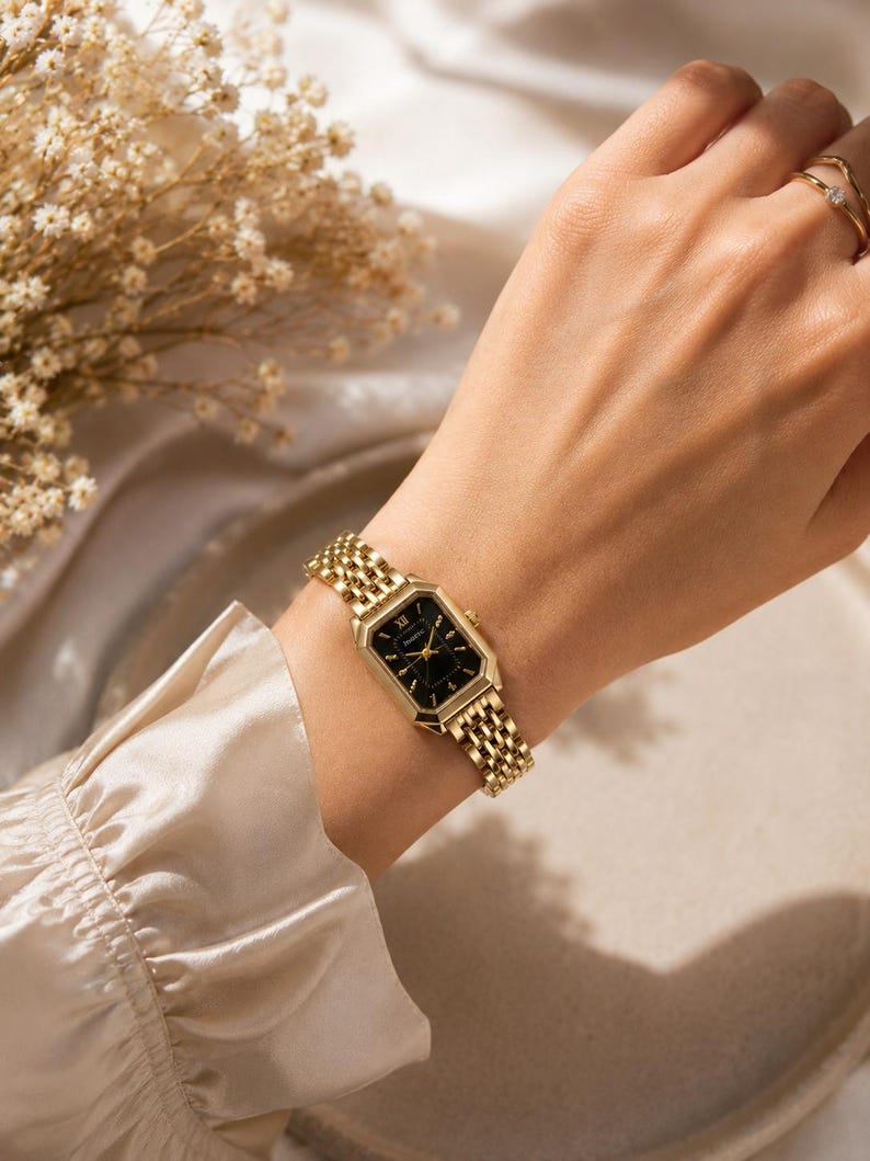 May include: A gold-toned wristwatch with a black face and Roman numeral hour markers is worn on a wrist. The watch has a rectangular face and a woven metal band. A gold ring is on the finger. The background includes a beige silk fabric and dried flowers.