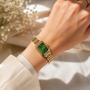May include: A gold-toned wristwatch with a rectangular green face and a matching gold band. The watch is worn on a wrist, showcasing its intricate design and elegant aesthetic. The watch face features Roman numerals.