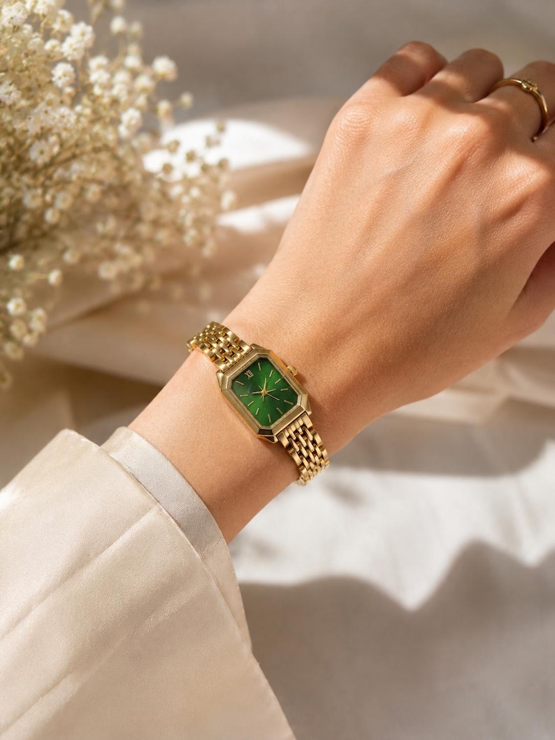 May include: A gold-toned wristwatch with a rectangular green face and a matching gold band. The watch is worn on a wrist, showcasing its intricate design and elegant aesthetic. The watch face features Roman numerals.