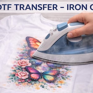 May include: A white t-shirt featuring a vibrant butterfly and floral design being ironed. The iron is blue and white. The image shows the DTF transfer process. The butterflies have orange, pink, purple, and yellow wings.