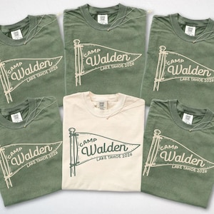 May include: Five short-sleeve crew neck t-shirts arranged in a group. Four shirts are moss green with cream-colored pennant graphics, and one center shirt is ivory with a dark green pennant graphic. Each features the text Camp Walden Lake Tahoe 2026.