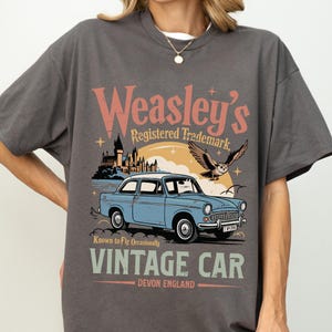 May include: Dark gray t-shirt featuring a vintage car graphic. The graphic includes the text "Weasley's Registered Trademark" and "Vintage Car Devon England". A light blue car is the focal point of the design, with an owl and castle in the background.