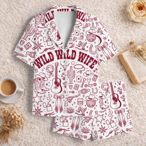 May include: A white short-sleeve button-down pajama shirt and matching shorts set featuring a repeating maroon western-themed print of guitars, cowboy boots, cacti, hats, and revolvers with the text WILD WILD WIFE printed across the chest.