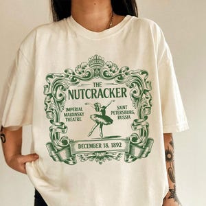 May include: Cream-colored t-shirt featuring a green graphic design for "The Nutcracker" ballet. The design includes text, a ballerina illustration, and the date December 18, 1892. The shirt is a relaxed fit.