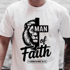 Könnte beinhalten: Weißes T-Shirt mit einem schwarzen Löwenkopf-Design und dem Text "MAN of Faith" sowie dem Bibelvers "1 Korinther 16:13". Die Grafik ist mittig auf dem T-Shirt platziert.