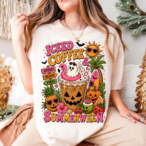 May include: A cream-colored t-shirt featuring a graphic print with the text "ICEED COFFEE & SUMMERWEEN". The design includes a jack-o'-lantern coffee cup, a ghost in a flamingo floatie, a sun with sunglasses, palm trees, and tropical fruit.