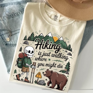 May include: Cream-colored t-shirt with a graphic featuring a skeleton hiker, mountains, trees, and a bear. The text reads "Hiking is just walking where you might die." A hiking-themed design.