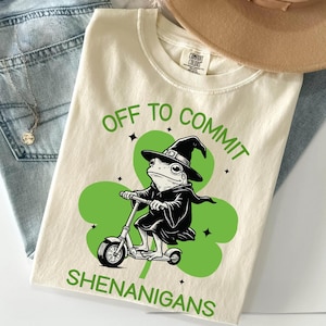 May include: Cream-colored t-shirt with a green graphic of a frog wearing a witch hat and cloak, riding a scooter on a green shamrock. The text "OFF TO COMMIT SHENANIGANS" is printed in green.