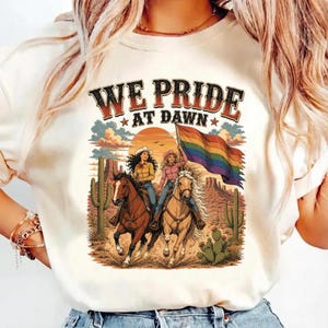 May include: Cream-colored t-shirt featuring a graphic of two women riding horses in a desert landscape, holding a rainbow flag. The text "WE PRIDE AT DAWN" is arched across the top of the design. Cacti and a sunset complete the design.