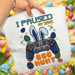 Op de afbeelding: Wit kussen met een pixelontwerp voor Pasen. Het ontwerp bevat een gamecontroller met konijnenoren, paaseieren en de tekst "I PAUSED MY GAME TO EGG HUNT" in groen en oranje.