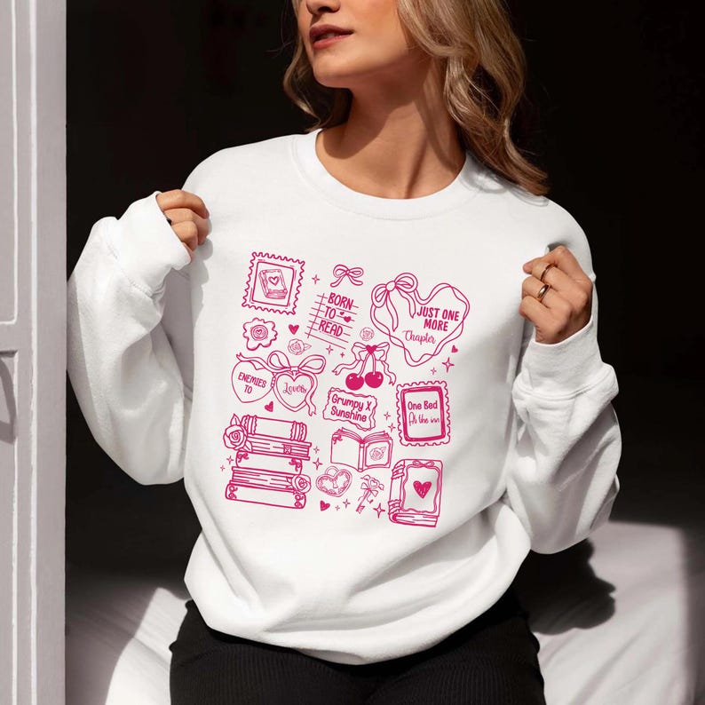Puede incluir: Sudadera blanca con un dise&ntilde;o gr&aacute;fico rosa que presenta ilustraciones y texto relacionados con libros. El dise&ntilde;o incluye corazones, libros y las frases "Born to Read" y "Just One More Chapter."