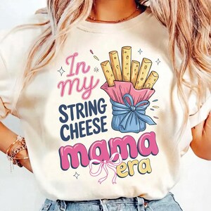 Könnte beinhalten: Cremefarbenes T-Shirt mit einem grafischen Design. Das Design zeigt den Text "In my string cheese mama era" in Pink, Blau und Gelb. Eine Cartoon-Illustration von Käsesnacks in einer rosa-blauen Verpackung ist ebenfalls enthalten.