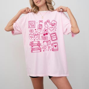 Puede incluir: Camiseta rosa claro con un dise&ntilde;o gr&aacute;fico rosa. El dise&ntilde;o incluye ilustraciones de libros, corazones y sellos, junto con texto como "Born to Read" y "Just One More Chapter".