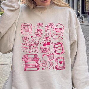 Puede incluir: Sudadera crema con un dise&ntilde;o gr&aacute;fico rosa que presenta ilustraciones y texto relacionados con libros. El dise&ntilde;o incluye libros apilados, corazones, lazos y frases como "Born to Read" y "Just One More Chapter."