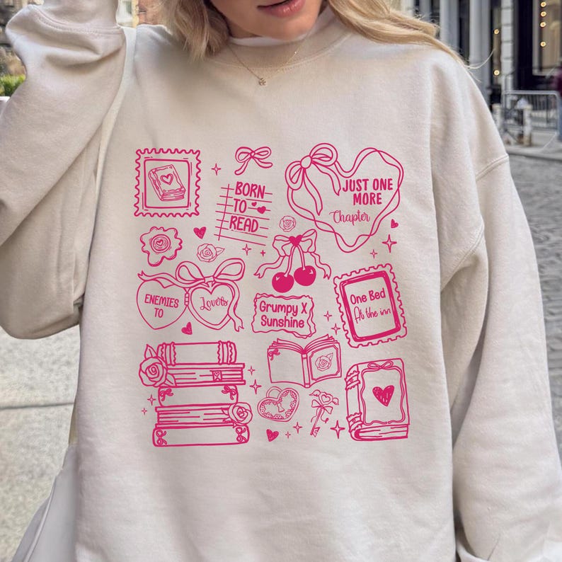 Puede incluir: Sudadera crema con un dise&ntilde;o gr&aacute;fico rosa que presenta ilustraciones y texto relacionados con libros. El dise&ntilde;o incluye libros apilados, corazones, lazos y frases como "Born to Read" y "Just One More Chapter."