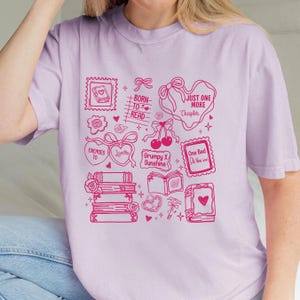 Puede incluir: Camiseta lila claro con un dise&ntilde;o gr&aacute;fico rosa de libros, corazones y texto como "Born to Read" y "Just One More Chapter". El dise&ntilde;o tiene una est&eacute;tica vintage.
