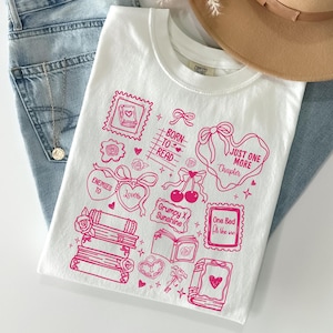 Puede incluir: Camiseta blanca con un dise&ntilde;o gr&aacute;fico rosa con ilustraciones tem&aacute;ticas de libros y texto. El dise&ntilde;o incluye corazones, lazos y las frases "Born to Read" y "Just One More Chapter."