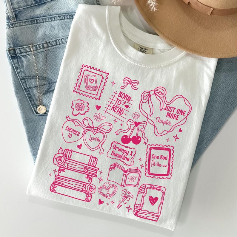 Puede incluir: Camiseta blanca con un dise&ntilde;o gr&aacute;fico rosa con ilustraciones tem&aacute;ticas de libros y texto. El dise&ntilde;o incluye corazones, lazos y las frases "Born to Read" y "Just One More Chapter."
