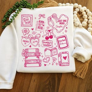 Puede incluir: Sudadera blanca con ilustraciones gr&aacute;ficas rosas de libros, corazones, lazos y texto. El texto incluye frases como "Born to Read", "Just One More Chapter" y "Grumpy X Sunshine". El dise&ntilde;o es una colecci&oacute;n de elementos tem&aacute;ticos de libros.