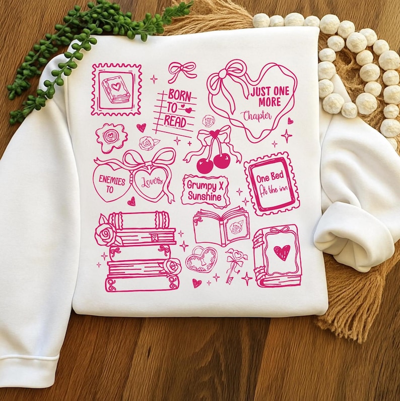 Puede incluir: Sudadera blanca con ilustraciones gr&aacute;ficas rosas de libros, corazones, lazos y texto. El texto incluye frases como "Born to Read", "Just One More Chapter" y "Grumpy X Sunshine". El dise&ntilde;o es una colecci&oacute;n de elementos tem&aacute;ticos de libros.