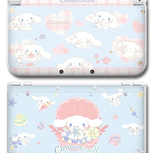 May include: A light blue Nintendo 3DS case featuring the character Cinnamoroll. The top half has a repeating pattern of Cinnamoroll and pink hearts. The bottom half has a large Cinnamoroll design inside a seashell, with the name "Cinnamoroll" printed below.