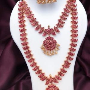 May include: A gold-toned jewelry set featuring a long necklace and a shorter necklace, both adorned with red stones in a paisley pattern and small white beads. The set includes matching bell-shaped jhumka earrings with red stones and white beads.