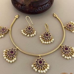 Puede incluir: Un collar y un par de pendientes a juego en tono dorado con piedras circulares de color rojo oscuro rodeadas de pequeñas cuentas blancas. El collar tiene un diseño de cadena flexible con cinco colgantes florales, mientras que los pendientes presentan un colgante a juego con cierre de gancho.