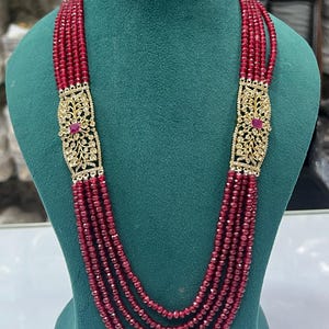 May include: A multi-strand necklace featuring deep red faceted beads arranged in four cascading layers. Two rectangular gold-toned metal connectors are positioned on each side, decorated with intricate filigree patterns and a central red stone.