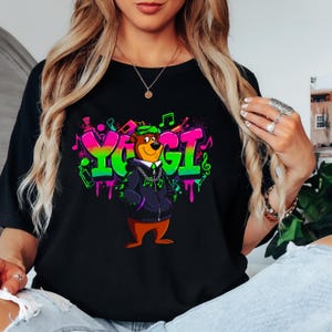 May include: Black t-shirt featuring a cartoon bear character with the word "YOGI" in graffiti style. The bear wears a green hat and a black jacket with a silver chain. Musical notes and colorful paint splatters surround the text.