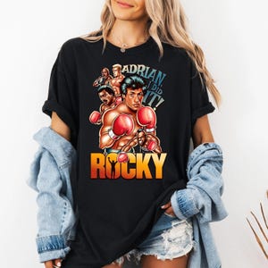 Op de afbeelding: Zwart t-shirt met een afbeelding van Rocky Balboa met bokshandschoenen, met de tekst "ADRIAN, I DID IT!" en "ROCKY" in oranje en geel. Het ontwerp bevat afbeeldingen van andere personages uit de film.
